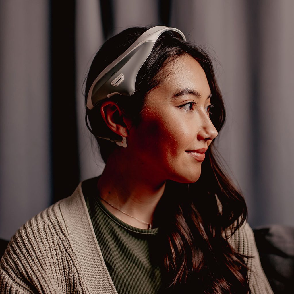 Side profile of a woman seated indoors, wearing the Modius Sleep neurostimulation headset over her forehead and behind her ears.